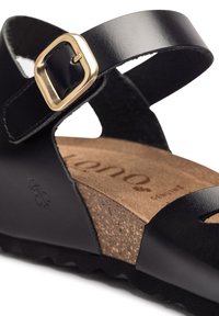 Sandale en cuir noir avec une boucle dorée, semelle en liège et semelle texturée. Présente un design minimaliste et un bout ouvert avec une sangle.