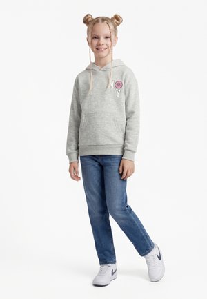 Mlada deklica stoji, oblečena v sivo kapucar z motivom krofa, modre kavbojke, bele superge in dveh spetih fig, nasmejana.