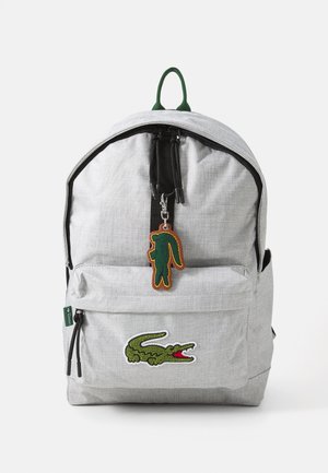Grau farbener Stoffrucksack mit einer Vordertasche, schwarzen Reißverschlüssen und einem grünen Krokodil-Logo. Enthält einen grünen Krokodil-Anhänger als Akzent.