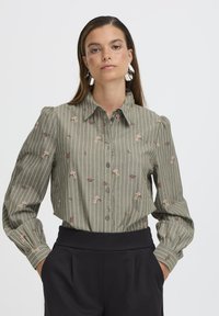 Camisa verde oliva de botones con rayas verticales blancas, bordado floral en rosa y rojo, y mangas largas abullonadas. Pantalones negros de cintura alta.