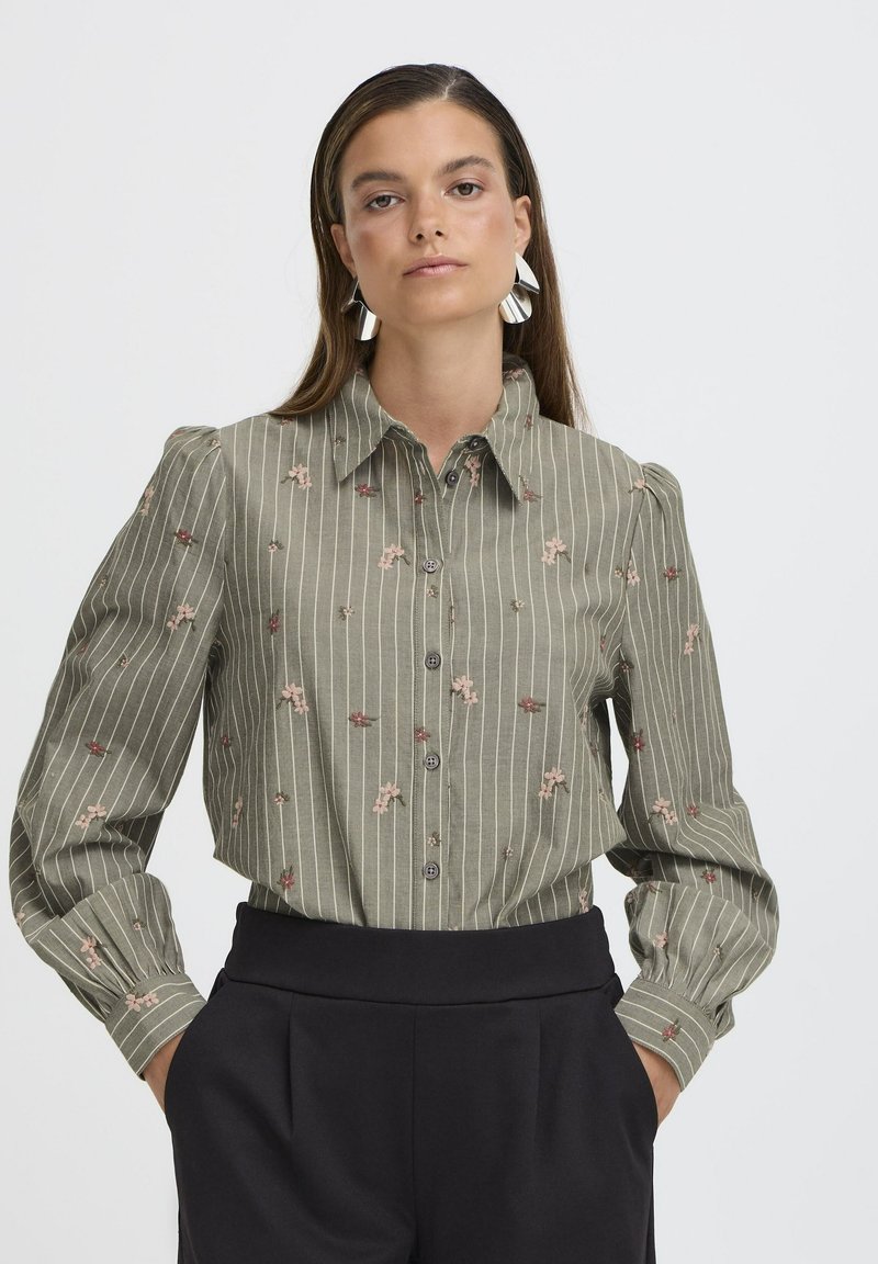 Camisa verde oliva de botones con rayas verticales blancas, bordado floral en rosa y rojo, y mangas largas abullonadas. Pantalones negros de cintura alta.