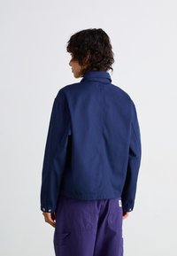 Veste bleu marine avec un col montant, coutures visibles et fermetures à pression au bas, associée à un pantalon cargo violet.