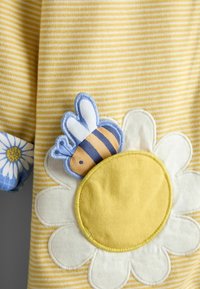 Geltonai baltos dryžuotos medžiagos su baltos ir geltonos ramunės aplikacija bei už gėlės dalinai pasislėpusia mėlynos ir geltonos bitės aplikacija.