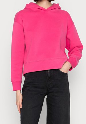 Persoon gekleed in een felroze hoodie en zwarte broek met hoge taille, één hand in de zak, bij de nek en bovenbenen bijgesneden.