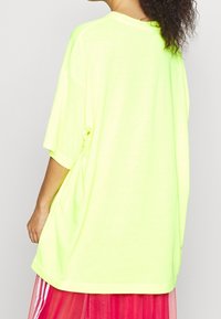 T-shirt oversize giallo neon con scollo tondo e maniche corte, mostrato da dietro, abbinato a una gonna rosa a strati.