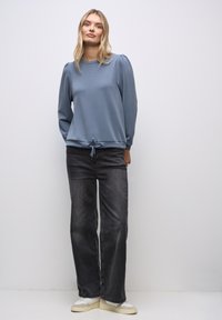 Sininen vetoketjupaita puffihihoilla ja säädettävällä nyörivedolla, yhdistettynä loseihin mustiin farkkuihin ja vaaleisiin tennareihin.