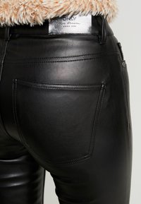 Černé kožené kalhoty vysokého pasu s detaily na zadních kapsách, hladká struktura a stříbrná značka na pase.