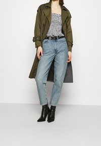 Trench-coat olive, haut à carreaux noir et blanc, jeans taille haute bleu clair, bottes noires à talons pointus.