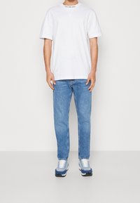 Bílý bavlněný tričko s kulatým výstřihem a černým logem, v kombinaci se světle modrými džínami a vícebarevnými sportovními botami.