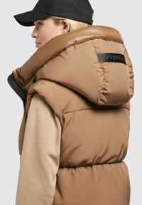 Gilet matelassé en marron clair avec un col haut, présentant des détails de logo noirs et une texture lisse, porté sur un haut à manches longues beige.
