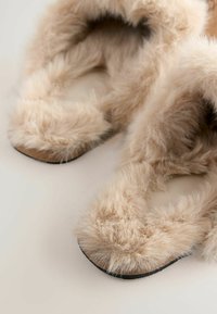 Chaussons en fausse fourrure de couleur beige clair avec une surface douce et texturée. Design à bout ouvert avec une semelle plate, comportant des détails en peluche autour des bords.