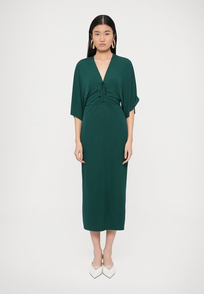 Grünes Midikleid mit V-Ausschnitt, Raffdetail an der Taille, weiten Ärmeln und glatter Textur. Kombiniert mit weißen spitzen High Heels.
