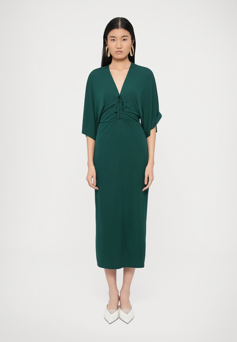 Πράσινο μίντι φόρεμα με V-λαιμόκοψη, λεπτομέρεια �σούρωσης στη μέση, φαρδιά μανίκια και λεία υφή. Φορεμένο με λευκές γόβες μυτερές.