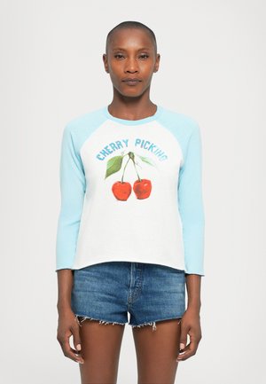 Mujer con la cabeza rapada que lleva una camisa de manga larga blanca y azul claro con un gráfico de cereza y pantalones cortos de mezclilla frente a un fondo blanco.