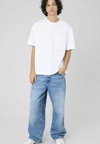T-shirt en coton blanc avec une poche sur la poitrine, associé à un jean en denim bleu clair à jambes larges. Le modèle se tient décontracté avec une main dans la poche.