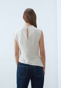 Haut en lin beige clair à manches courtes, détail œillet dans le dos, coupe ajustée et texture lisse, associé à un pantalon en denim foncé.