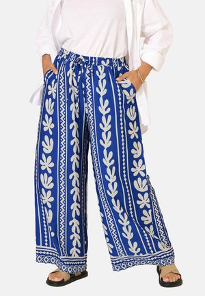 Pantalon bleu à jambes larges avec motifs floraux et géométriques blancs verticaux, taille élastique, associé à un haut blanc et une chemise blanche ouverte.