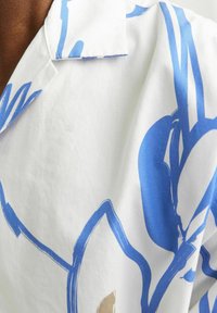 Camisa de manga corta en tejido blanco, con un gran estampado floral azul y detalles en marrón, y un cuello con muesca. Textura suave, diseño casual.