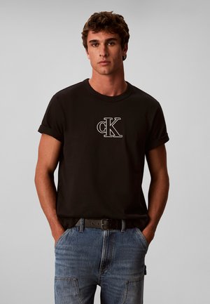 Junger Mann mit Händen in den Taschen trägt ein schwarzes T-Shirt mit aufgerollten Ärmeln und weißem CK-Logo, kombiniert mit blauen Jeans und schwarzem Gürtel.