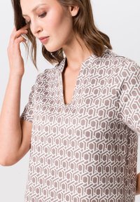 Blouse à manches courtes en beige clair avec un motif géométrique, dotée d'un décolleté en V et d'une texture lisse. Soulignée par des détails subtils.