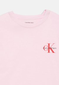 Gaiši rozā kokvilnas T-krekls ar apaļu apkakli. Uz kreisā krūts laukuma ir melns un sarkans "Calvin Klein" prints. Mīksta tekstūra.