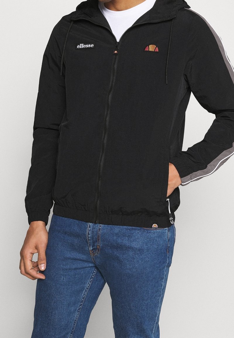 Homme portant une veste à capuche noire zippée avec une bande blanche et grise sur la manche, un jean bleu et une chemise blanche en dessous.