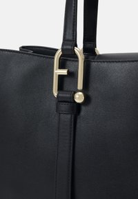 Sac en cuir noir avec un fermoir en métal doré arborant un logo stylisé, une surface texturée et des sangles plates pour le transport.