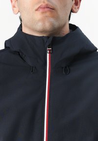 Námořnická kapuce s předním zipem a červenobílými akcenty. Lehký materiál s hladkou texturou a nastavitelnou kapucí.