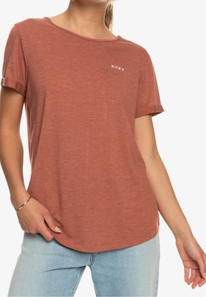 Kvinde iført en rustfarvet T-shirt med korte ærmer og "ROXY"-logo på brystet samt lyseblå jeans mod en hvid baggrund.
