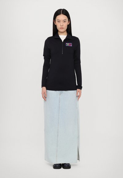 Black long-sleeve zip-up top with a colorful logo patch, paired with a light blue denim maxi skirt featuring a side slit.