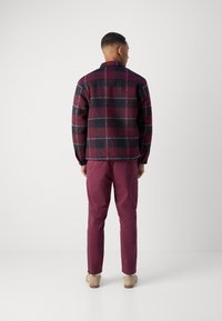 Veste en polaire à carreaux de couleur rouge foncé et noir, avec un col à boutons et des poches latérales, associée à un pantalon bordeaux foncé et des chaussures claires.