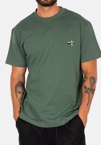 T-shirt in cotone verde oliva con collo tondo e grafica di una macchina fotografica nera sulla tasca sinistra del petto. Maniche corte, vestibilità rilassata.