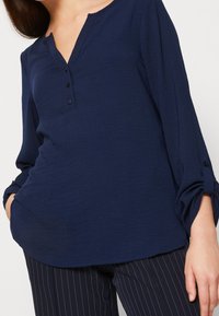 Femme portant un chemisier bleu marine à manches longues retroussées et un pantalon bleu marine à fines rayures, avec une main dans la poche.