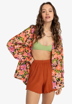 Blommönstrad, oversized skjorta i rosa och orange på en svart bakgrund, matchad med grön ribbad bikinitopp och rostfärgade shorts.