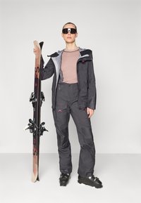 Ski outfit featuring a dark grey jacket with a light grey hood, pink patterned top, grey ski pants, and black ski boots, holding skis.