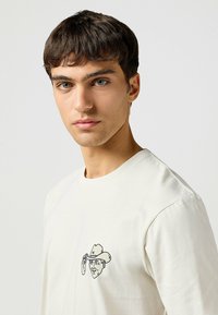 White cotton t-shirt featuring a printed graphic of a man wearing a cowboy hat, located on the left chest area. Crew neck and relaxed fit.