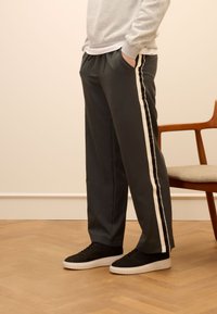 Graue Sporthosen mit weißen Seitenstreifen, ausgestattet mit einem Kordelzugbund und seitlichen Taschen, kombiniert mit schwarzen Sneakers.