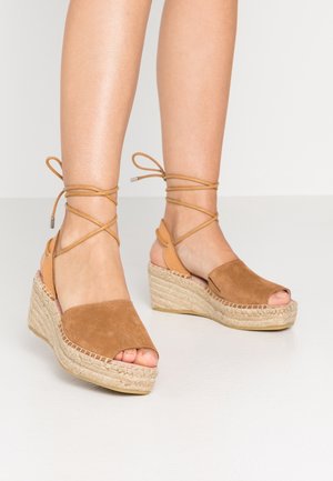 Sandales compensées en daim marron avec orteils ouverts et liens à nouer autour de la cheville, portées sur jambes nues contre un fond neutre.