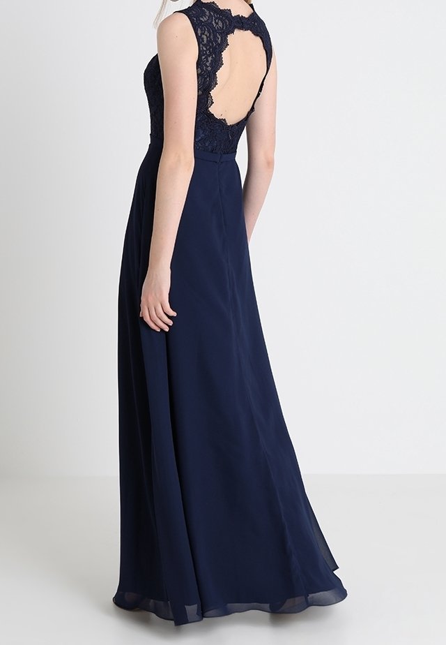 Femme portant une robe longue bleu marine avec un top en dentelle sans manches et un dos ouvert, debout avec sa main gauche reposant à ses côtés.