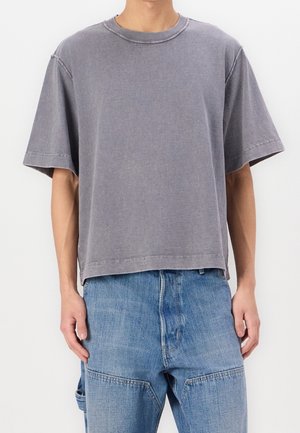 Man wearing a faded gray oversized short-sleeve t-shirt and light blue patchwork denim jeans standing against a plain white background.
