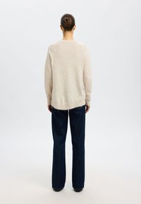 Pulover fuzzy, de culoare bej deschis, cu mâneci lungi, guler rotund și spate mai lung. Însoțit de blugi evazati de culoare albastru închis și pantofi negri.