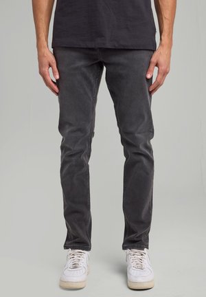 Hombre con vaqueros ajustados negros descoloridos y zapatillas blancas, de pie contra un fondo liso gris claro.