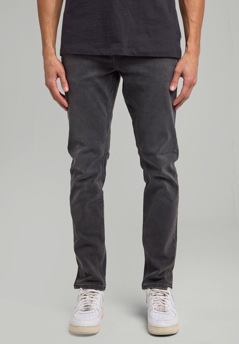 Mann trägt ausgewaschene schwarze Slim-Fit-Jeans und weiße Sneaker, steht vor einem schlichten hellgrauen Hintergrund.