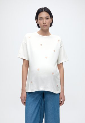 Γυναίκα με κοντά μαύρα μαλλιά που φοράει λευκό φαρδύ T-shirt με μικρά ροζ τριανταφυλλάκια και μπλε τζιν παντελόνι φαρδιάς γραμμής, στέκεται μπροστά σε λευκό φόντο.