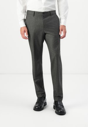 Man in een grijze, op maat gemaakte broek, een wit shirt en zwarte instapschoenen, staande tegen een plain witte achtergrond.