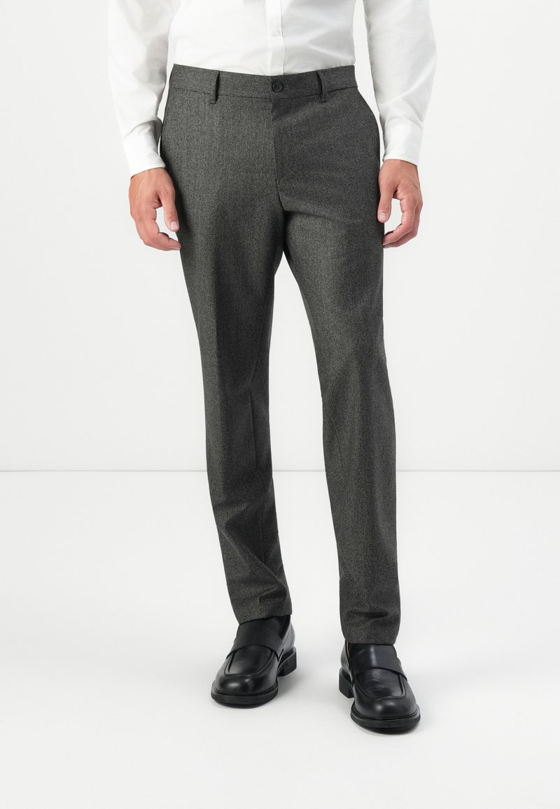 Homme portant un pantalon gris sur mesure, une chemise blanche et des mocassins noirs, se tenant devant un fond blanc uni.
