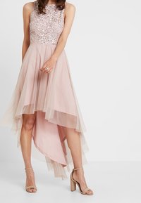 Jasnoróżowa sukienka z cekinowym gorsetem i warstwową tiulową spódnicą, z asymetrycznym dołem. Połączona z beżowymi sandałami na słupku.
