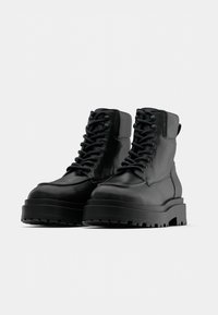 Černé kožené kotníkové boty s kulatou špičkou, silnou platformou a pevným šněrováním. Zahrnují polstrovaný límec a texturovanou podrážku s gripem.