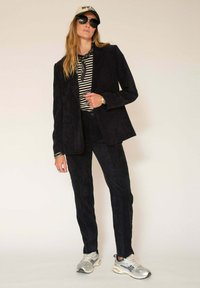 Blazer en velours côtelé noir et pantalon assorti, porté avec un haut rayé. Les accessoires comprennent des lunettes de soleil et une casquette beige. Baskets argentées.