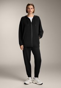 Felpa nera con zip, con lacci e tasche frontali, abbinata a pantaloni della tuta neri a gamba stretta e scarpe sportive grigie. Tessuto morbido.